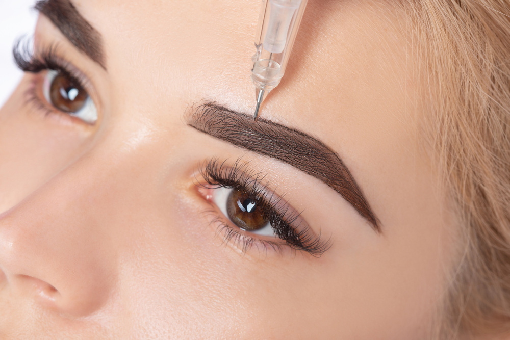 Permanent make-up for eyebrows of beautiful blonde woman in beauty salon. Closeup beautician doing eyebrows tattooing.
